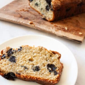 A slice of gluten free blueberry bread is served on a white plate with the loaf in the background.