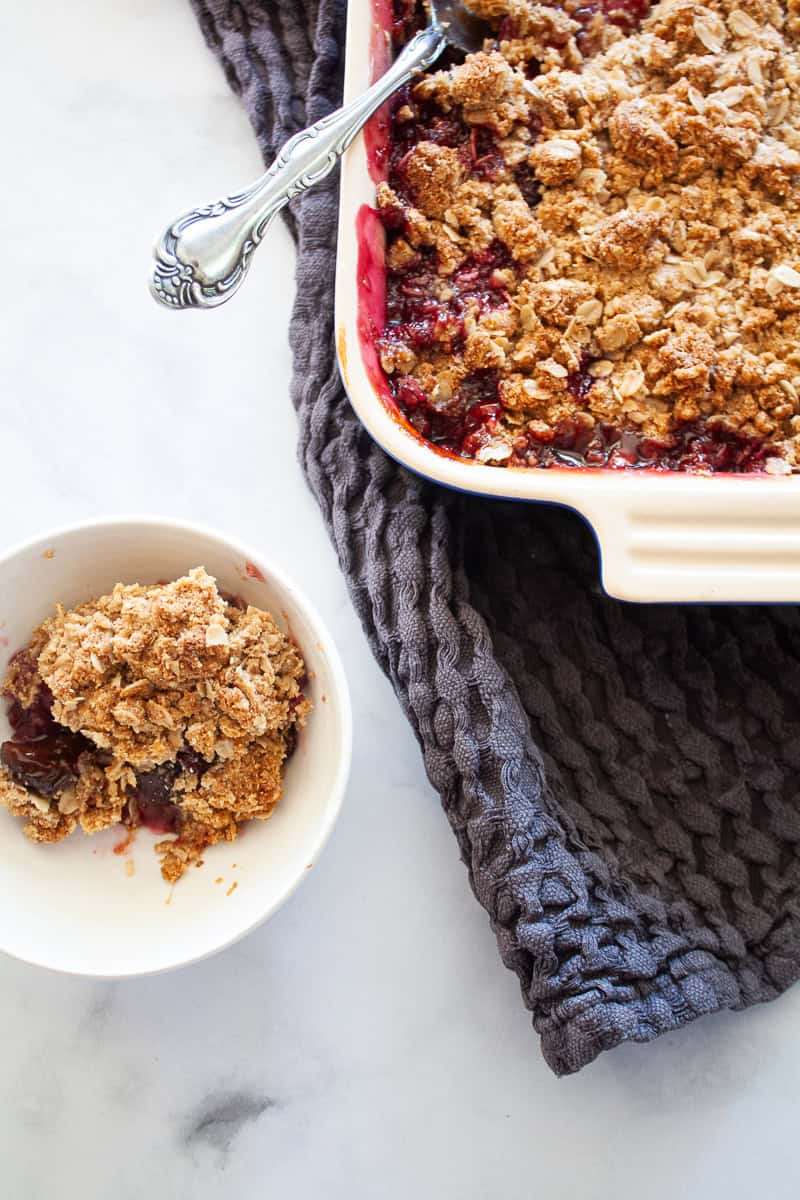 A bowl of gluten free cherry crisp in a white bowl to the left of a pa.n of crisp.