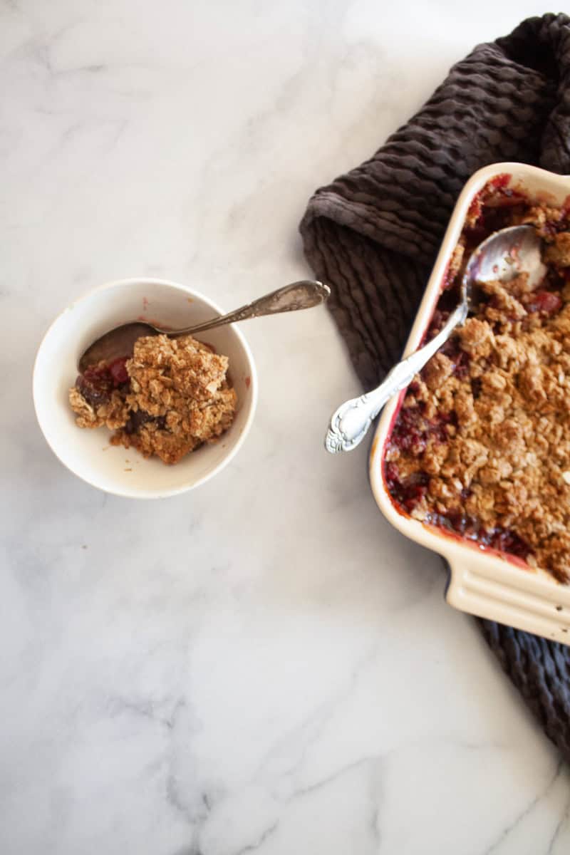 A bowl of cherry crisp with a spoon next to a pan of crisp.