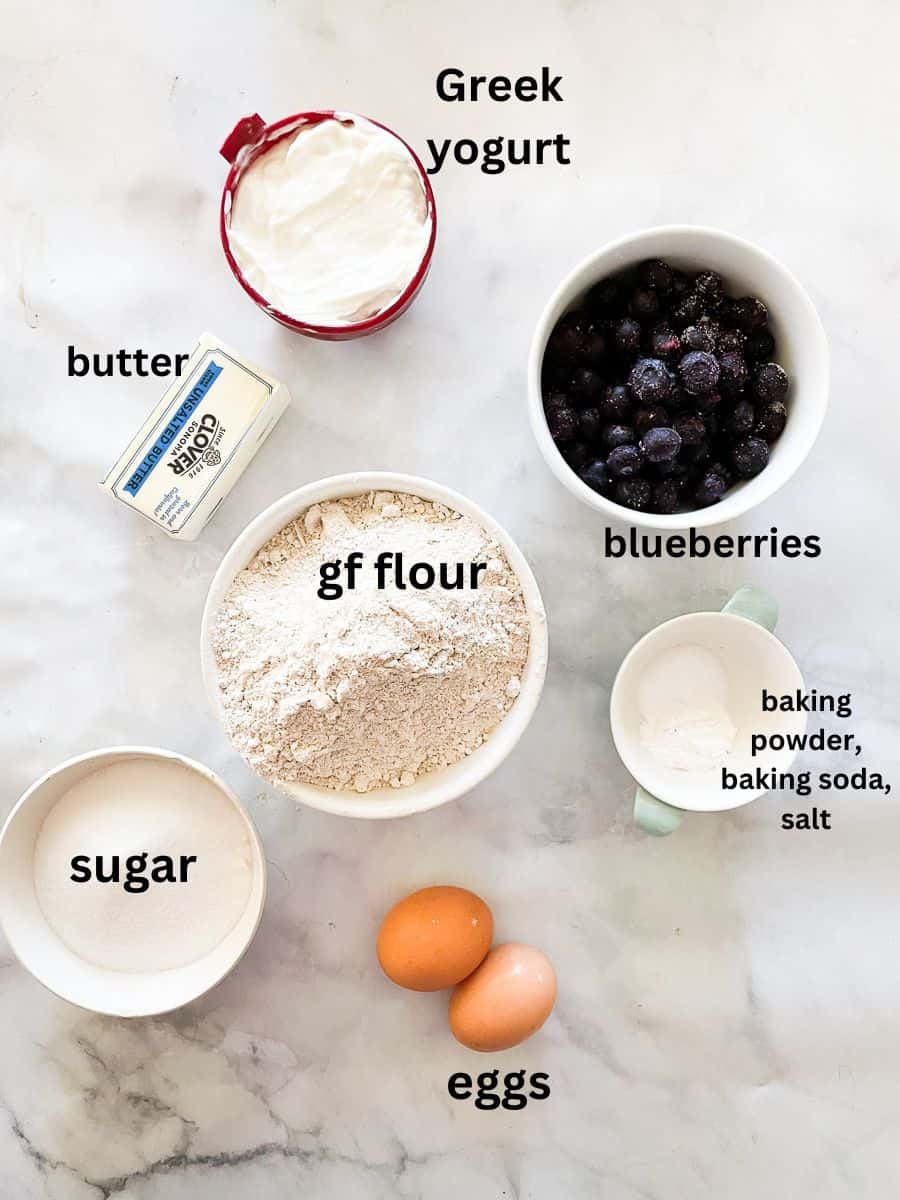 Ingredients for blueberry bread are shown labeled and portioned out: blueberries, gluten free flour, Greek yogurt, butter, baking powder, baking soda, salt, sugar, eggs.