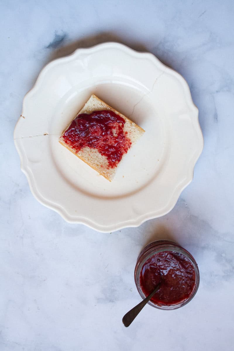 A jar of honey strawberry jam next to a piece of bread with jam.
