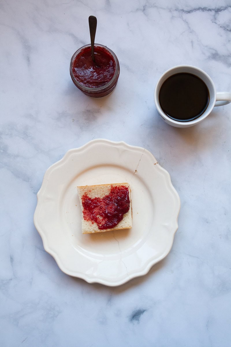A plate with bread and strawberry jam, a jar of jam, and a cup of coffee.