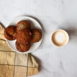 A latte next to a plate of gluten free strawberry banana muffins and a yellow towel.