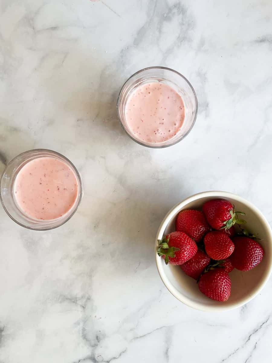 A bowl of strawberries alongside two glasses of banana strawberry smoothie.