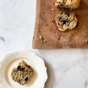 A slice of gluten free blueberry banana bread on a plate with the bread on a cutting board next to it.