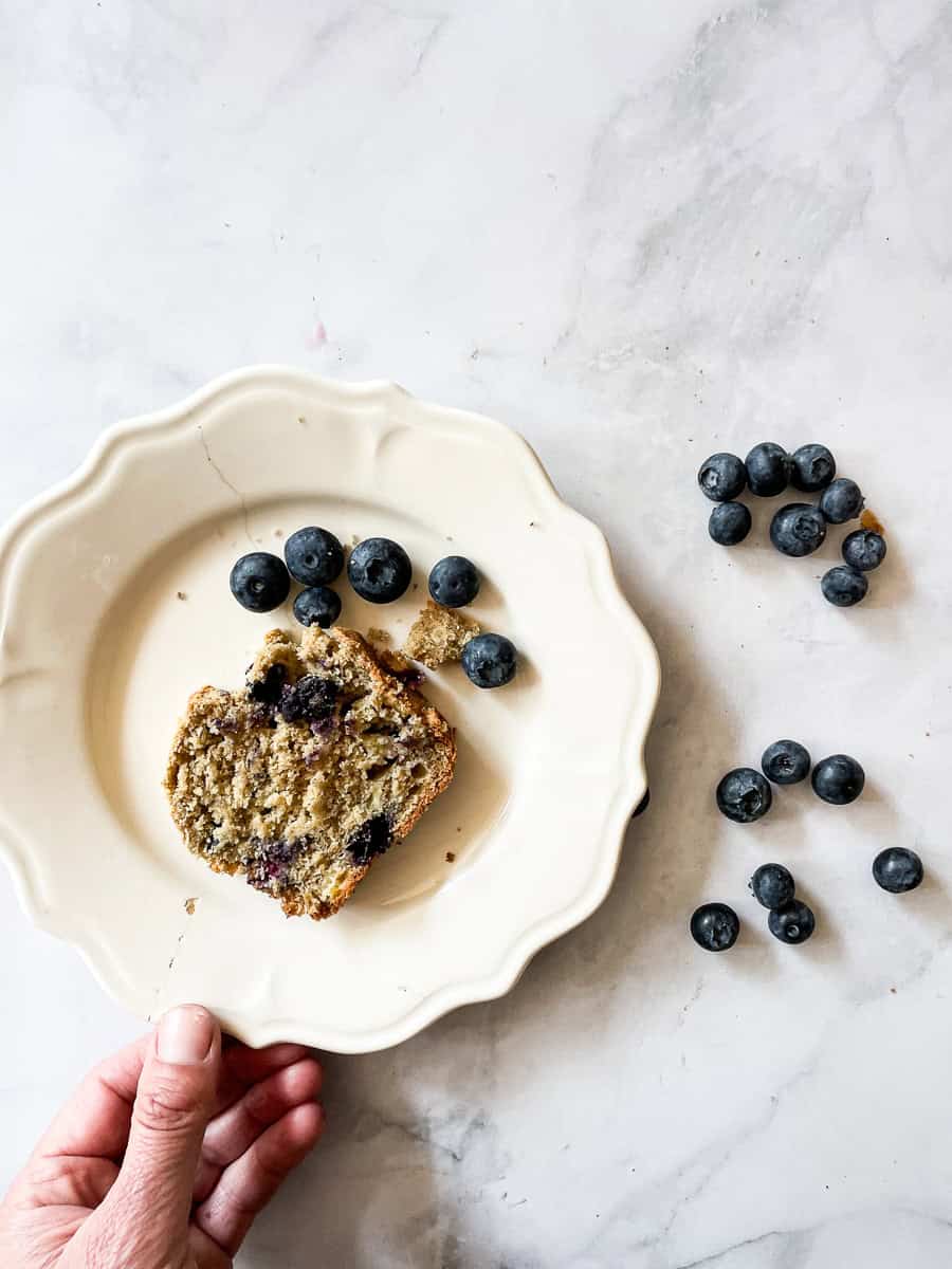 A hand touches a white plate with a slice of gluten free blueberry banana bread.