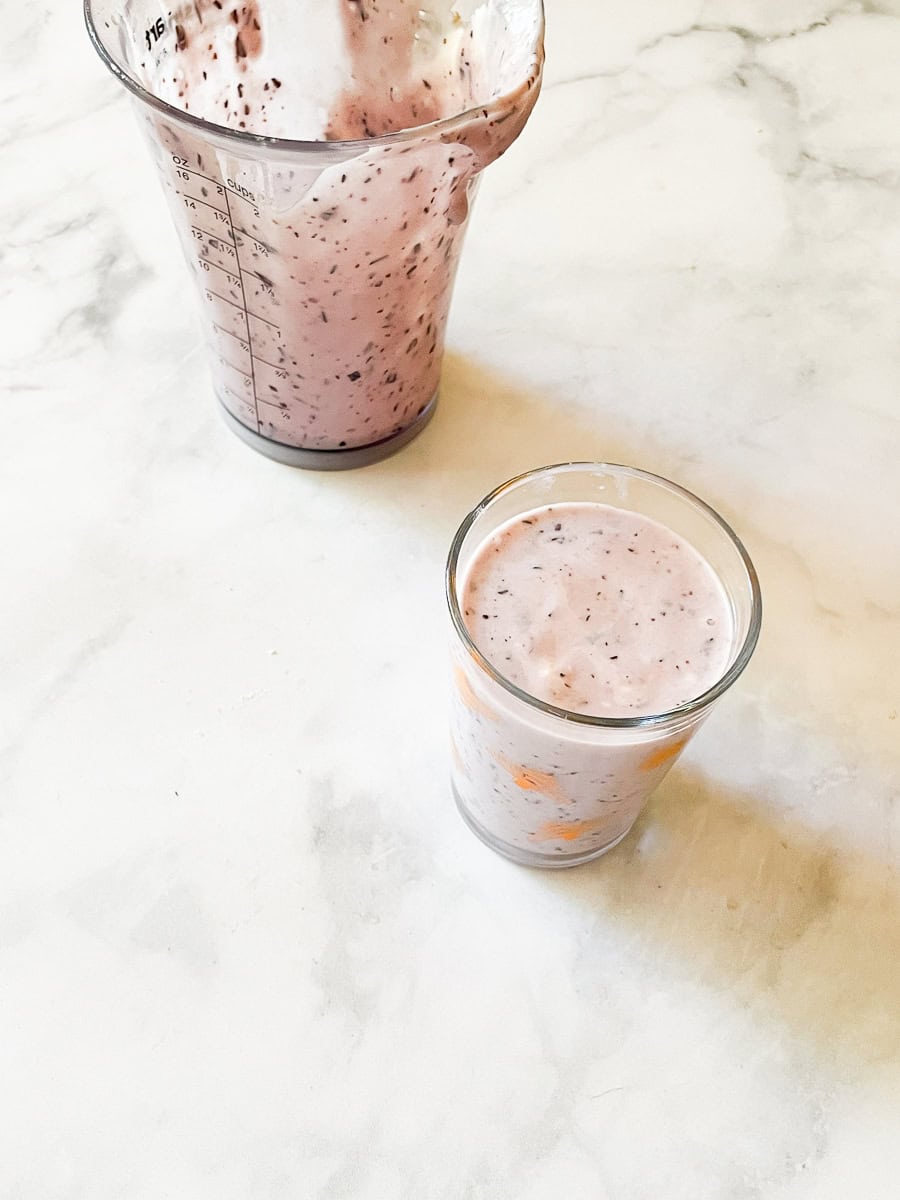 An empty pitcher of blueberry banana smoothie with a glass of smoothie in front of it.