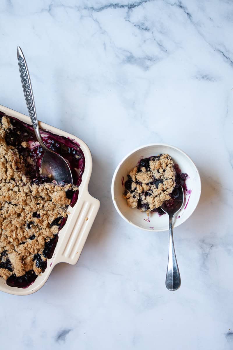 A bowl of blueberry crisp to the right of a pan of crisp.