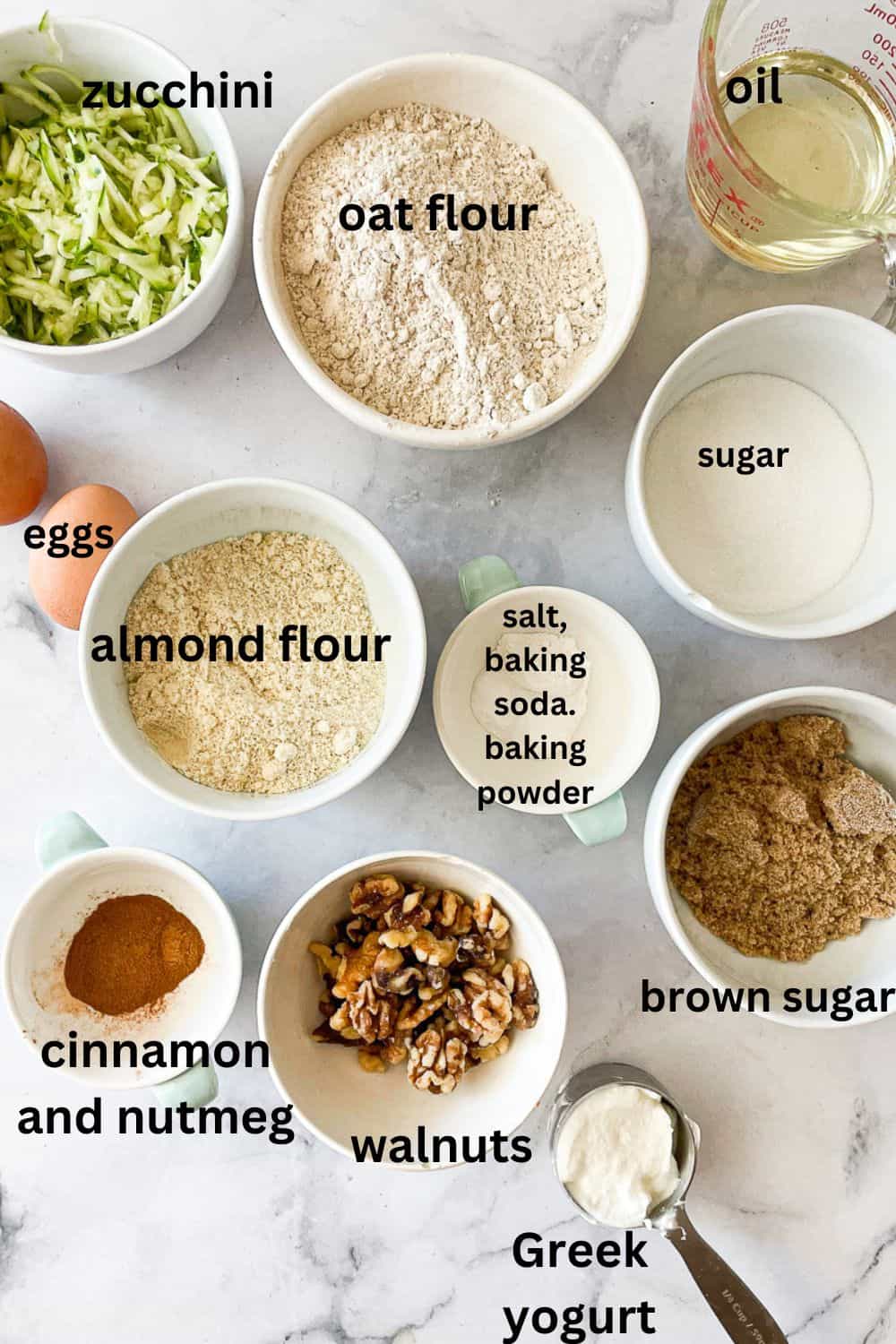 Labeled ingredients for gluten-free zucchini bread: shredded zucchini, brown sugar, sugar, baking soda, baking powder, salt, nutmeg, cinnamon, almond flour, oat flour, oil, eggs, Greek yogurt.