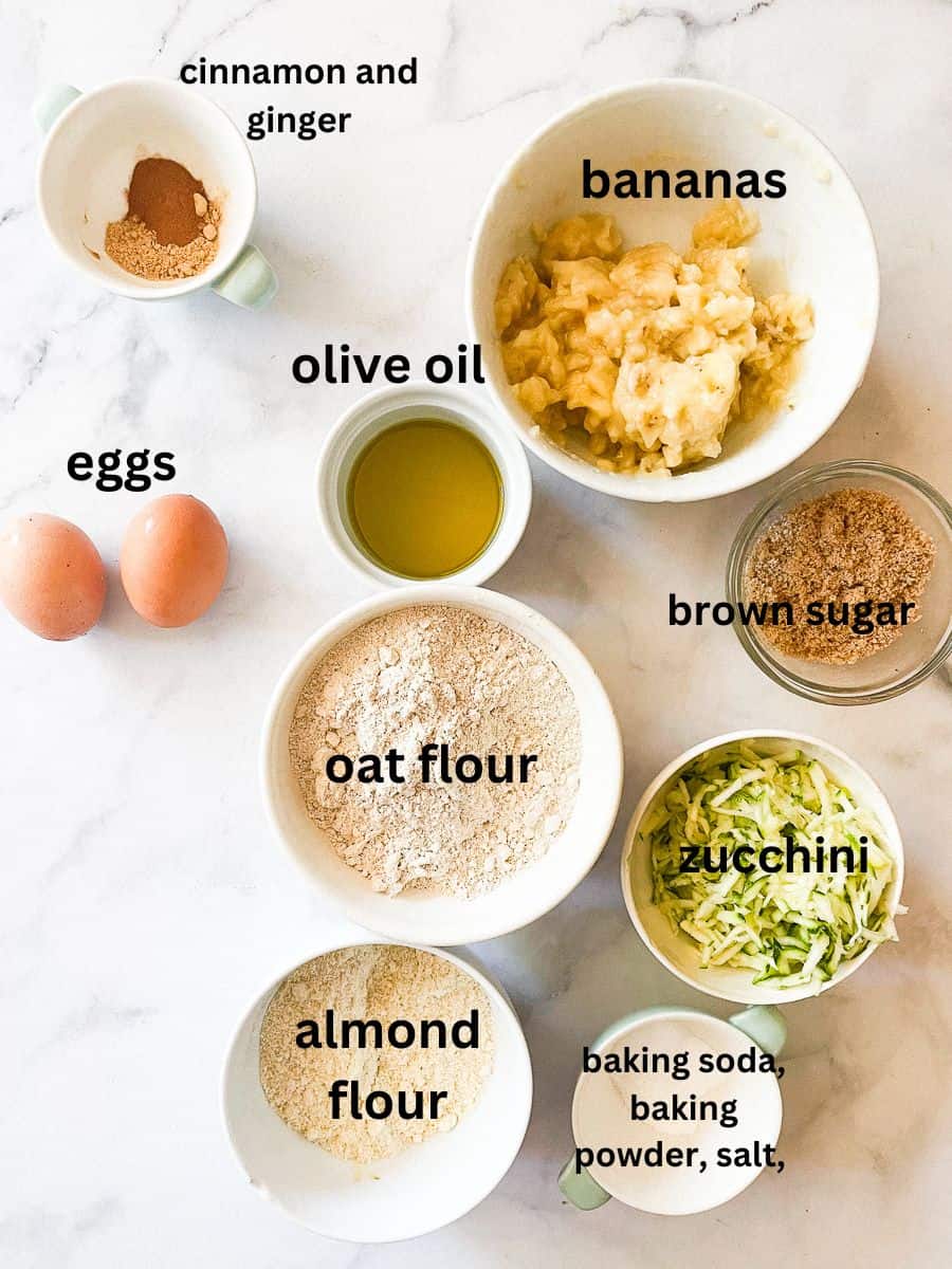 Labeled ingredients for gluten free banana zucchini muffins: almond flour, oat flour, olive oil, brown sugar, zucchini, eggs, ginger, cinnamon, baking powder, baking soda, and salt.