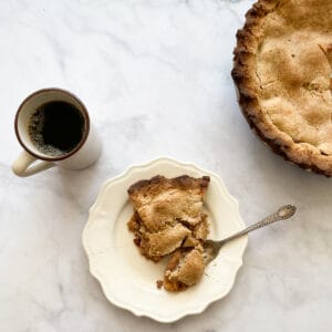 A piece of gluten free Gravenstein apple pie on a plate with the pie to the right and a cup of black coffee to the left.