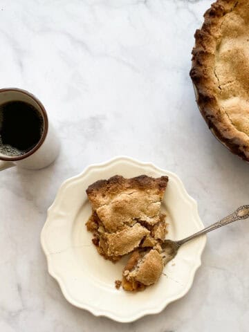 A piece of gluten free Gravenstein apple pie on a plate with the pie to the right and a cup of black coffee to the left.