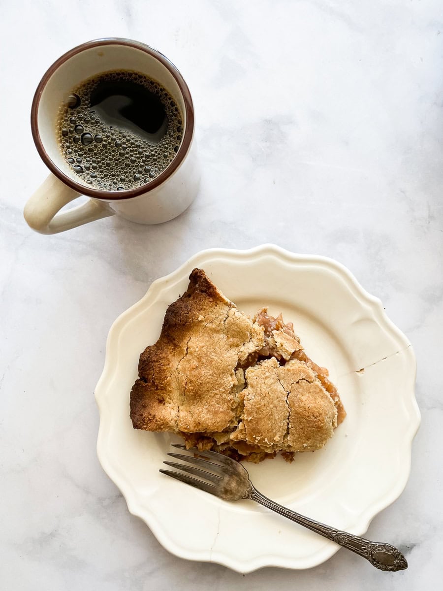 A cup of black coffee is shown to the left of a plate with a slice of gluten free Gravenstein apple pie and a fork.