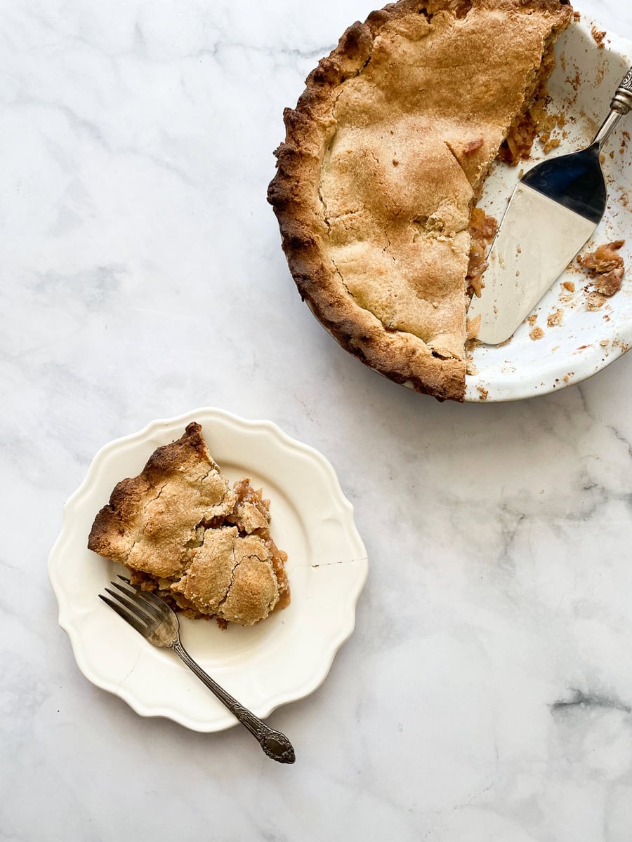 A slice of apple pie on a plate with the pie to the right.