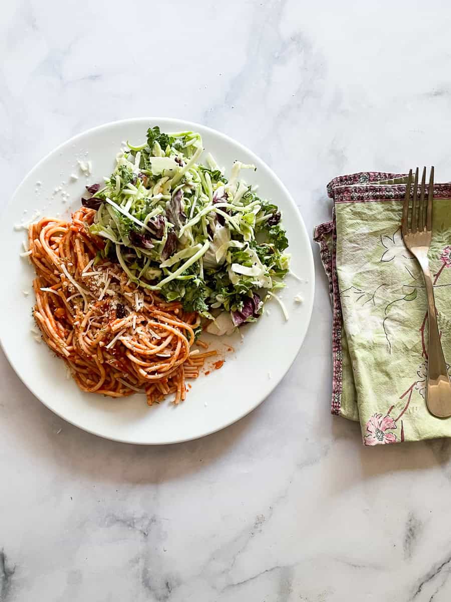 A plate of spaghetti with simple tomato sauce, salad, with a napkin and fork to the right.