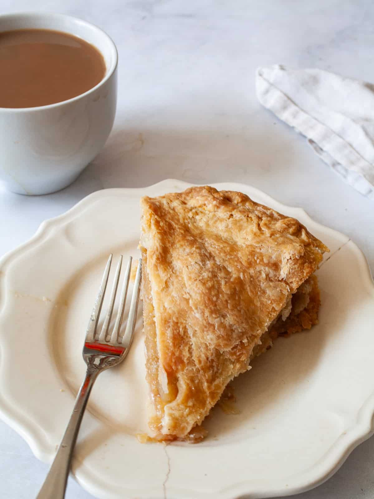 Fold tart-sweet apples into a flaky gluten free crust in this luscious gluten free Gravenstein apple pie recipe. A cup of coffee next to a slice of gluten free apple pie on a plate.