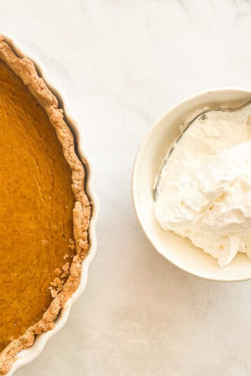 A bowl of whipped cream with a spoon next to a pumpkin pie from scratch.