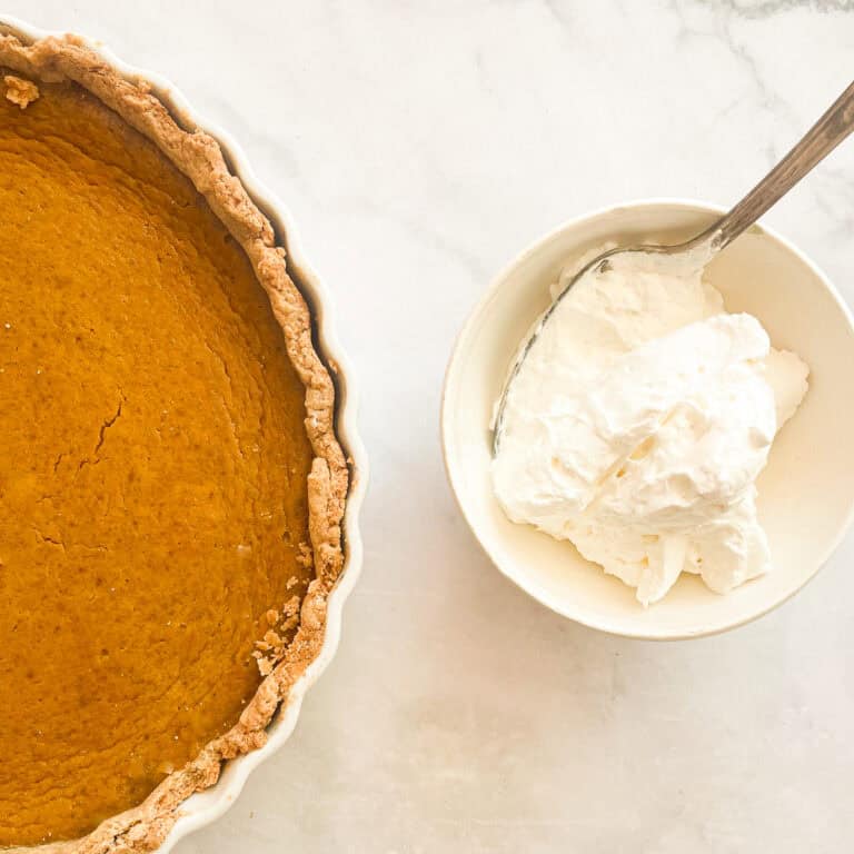 A bowl of whipped cream with a spoon next to a pumpkin pie from scratch.