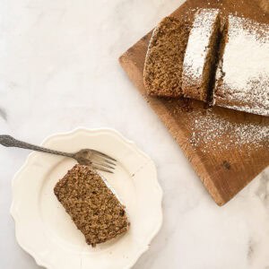 A slice of gingerbread on a plate with a fork and the loaf next to it.