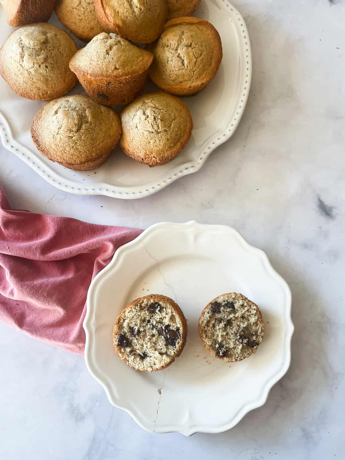 A chocolate chip muffin cut in half on a plate and a plate of muffins with a red napkin.