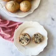 A chocolate chip muffin cut in half on a plate and a plate of muffins with a red napkin.