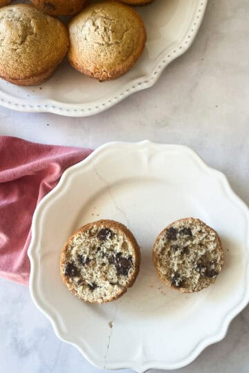 A chocolate chip muffin cut in half on a plate and a plate of muffins with a red napkin.