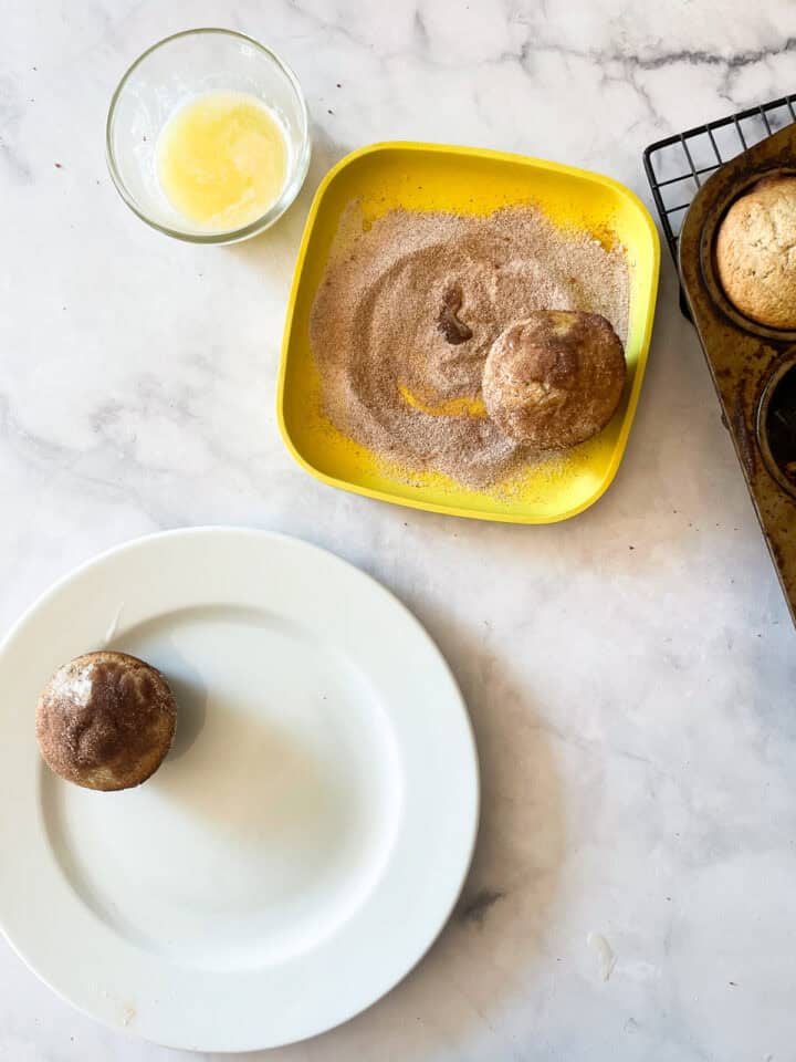 A donut muffin is dipped in cinnamon sugar and a muffin is on a plate.