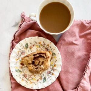 A cinnamon roll on a plate on a napkin with a cup of coffee.