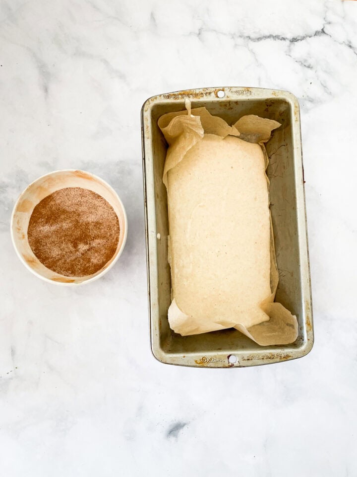 Cinnamon bread batter in a loaf pan with cinnamon sugar next to it.