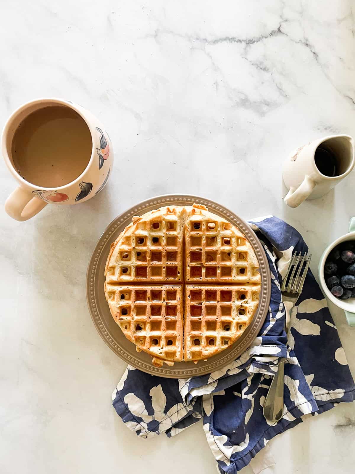 Waffles on a plate with a coffee cup and a pitcher of syrup next to them.