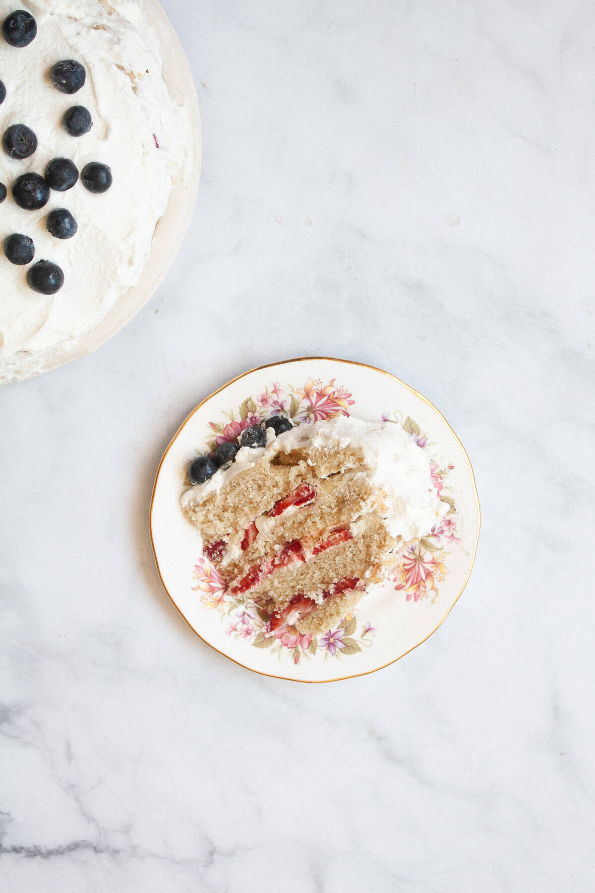 A gluten free berries and cream cake is layered with sweet whipped cream, sliced strawberries, and juicy blueberries. A berries and cream cake on a plate.