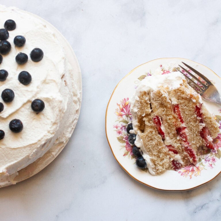 A slice of berries and cream cake on a plate with a fork next to the cake.