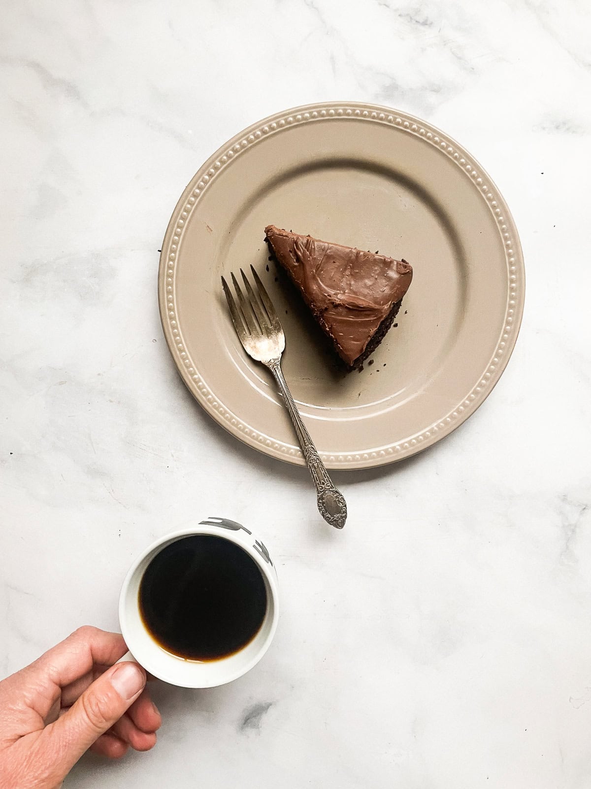 A slice of chocolate teff cake on a plate and a hand with a coffee cup.