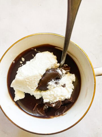 A tea cup of gluten free chocolate pudding and whipped cream with a spoon.