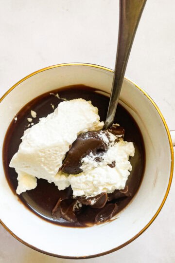 A tea cup of gluten free chocolate pudding and whipped cream with a spoon.
