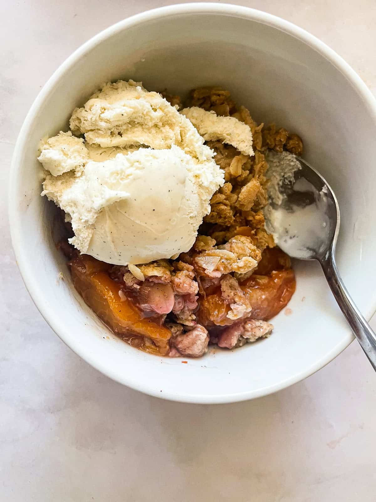 This gluten-free peach crisp stars juicy fresh peaches tucked under a rich toasted oat topping. Must be served with vanilla ice cream! A scoop of vanilla ice cream melts into a bowl of gluten-free peach crisp.