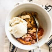 A scoop of vanilla ice cream melts into a bowl of gluten-free peach crisp.