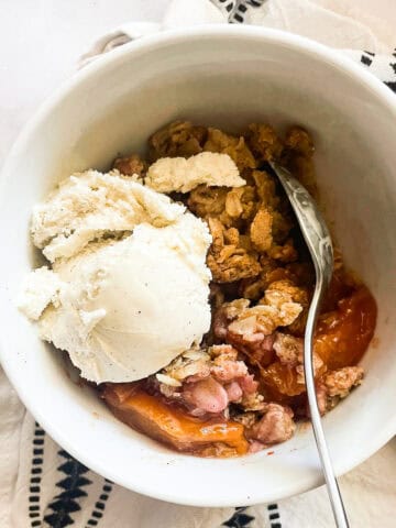 A scoop of vanilla ice cream melts into a bowl of gluten-free peach crisp.