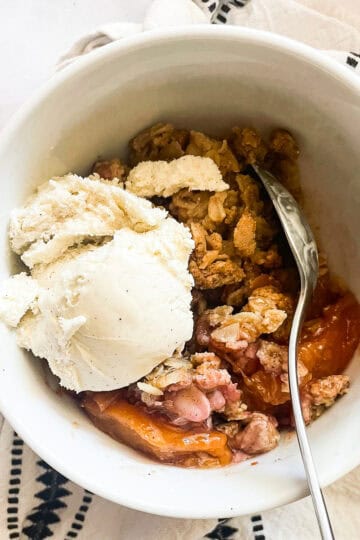 A scoop of vanilla ice cream melts into a bowl of gluten-free peach crisp.