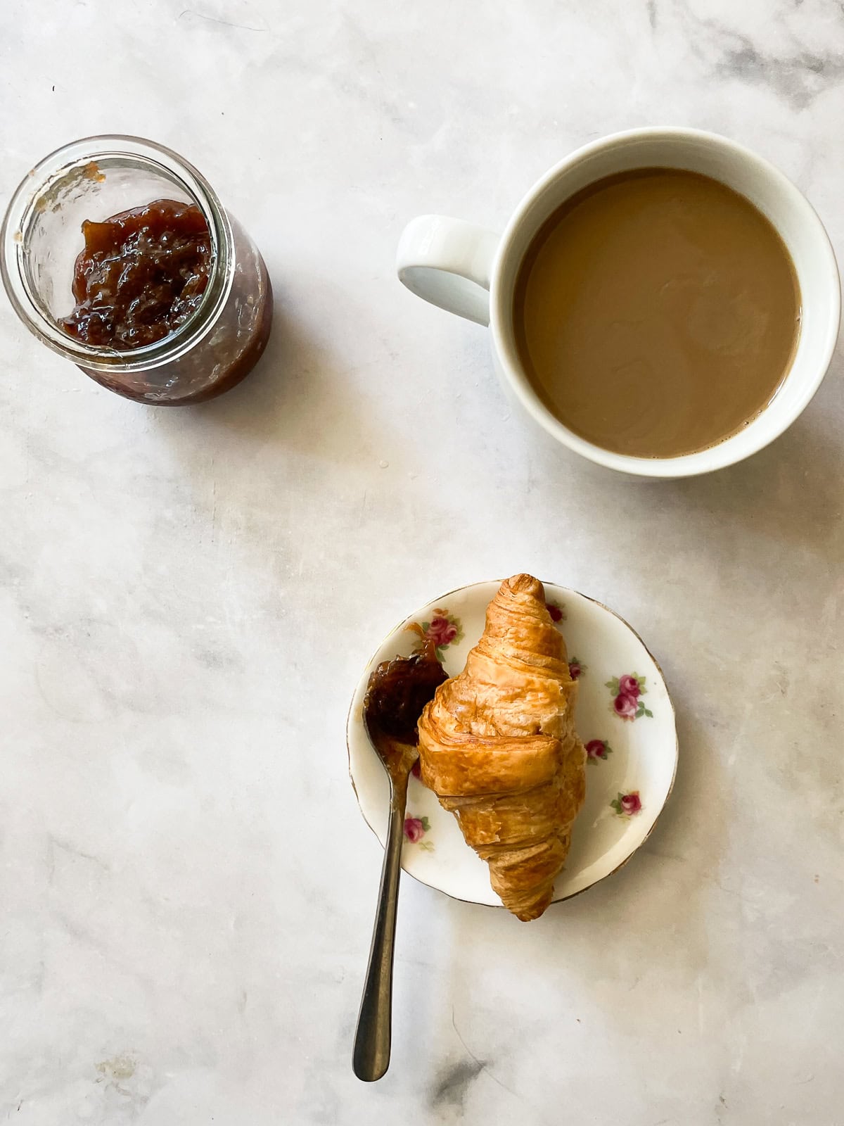 A mini croissant on a plate with a cup of coffee and a jar of strawberry & rhubarb jam.