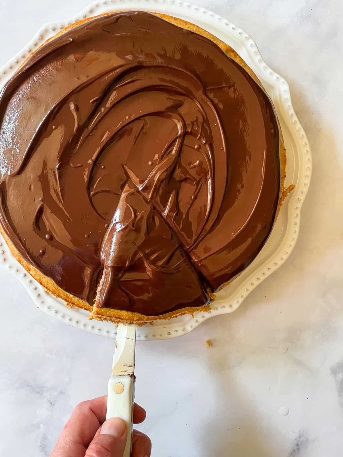 A lusciously moist, spice-kissed banana cake with chocolate frosting is the simplest recipe that always manages to impress! A hand uses a knife to cut a piece of banana cake with chocolate frosting.