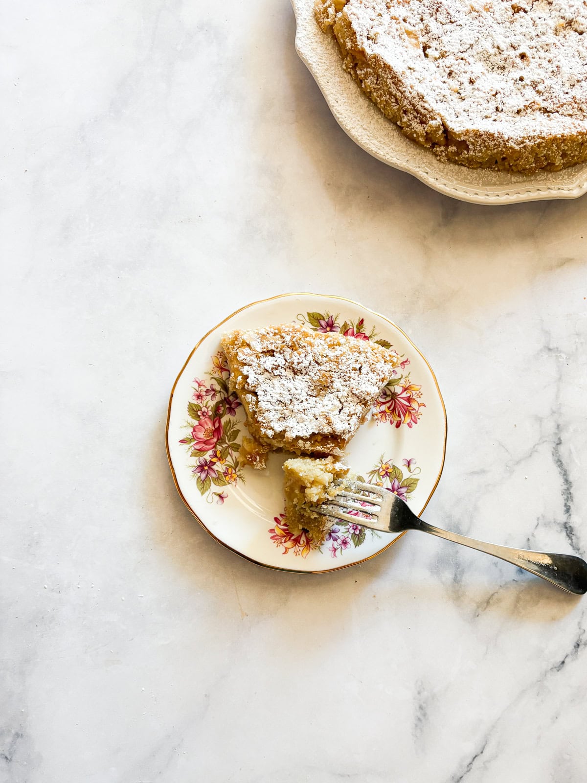 This buttery and tender gluten free French apple cake is inspired by a classic recipe and will almost literally melt in your mouth. A fork holds a bite of french apple cake next to a slice on a cake.