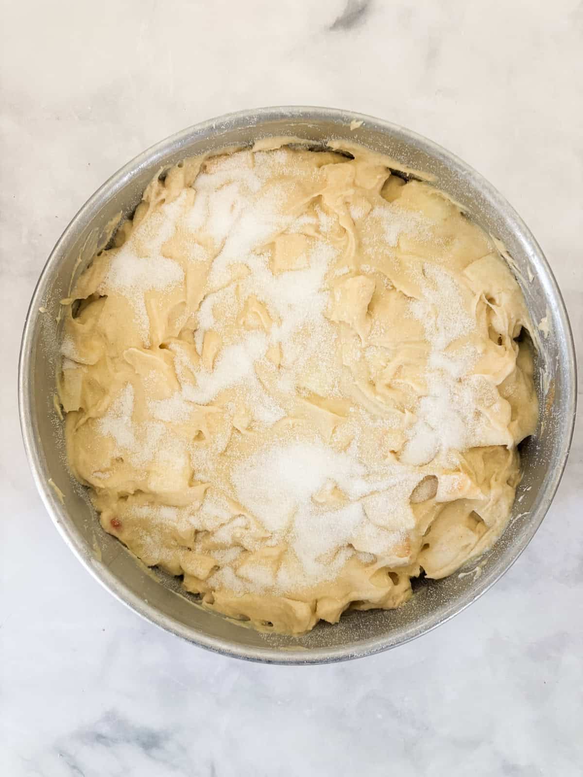 This buttery and tender gluten free French apple cake is inspired by a classic recipe and will almost literally melt in your mouth. The batter for french apple cake in a cake pan topped with granulated sugar.