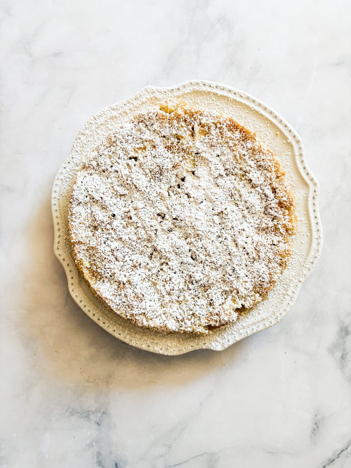 This buttery and tender gluten free French apple cake is inspired by a classic recipe and will almost literally melt in your mouth. A gluten free french apple cake dusted with powdered sugar on a white plate.