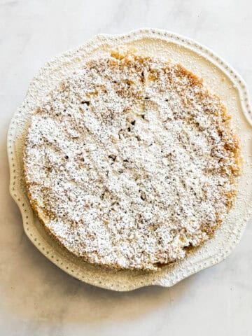A gluten free french apple cake dusted with powdered sugar on a white plate.