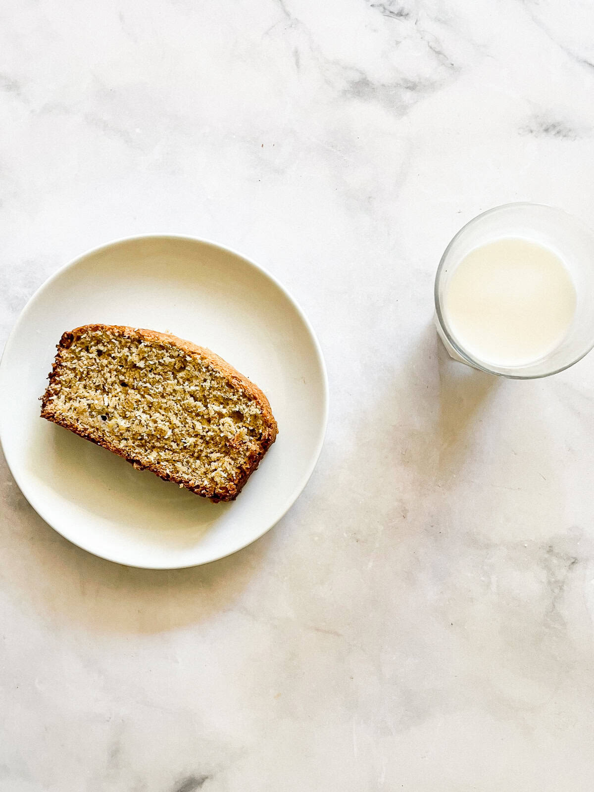 This recipe for gluten free coconut banana bread produces a beautiful loaf with a dense, moist crumb that's swirled with shredded coconut. A slice of gluten free coconut banana bread on a plate next to a glass of milk.