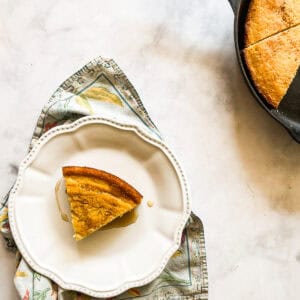 A slice of gluten free maple cornbread on a plate next to a pan of cornbread.