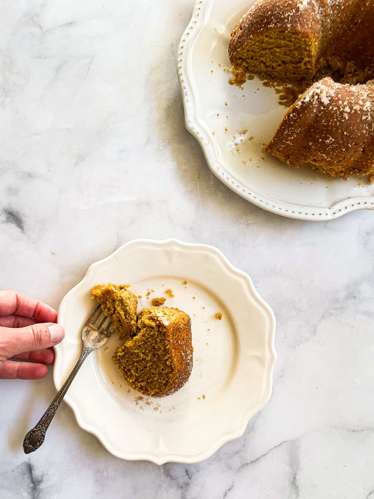 A soft and sumptuous gluten free pumpkin donut cake is bathed in cinnamon-sugar for a fabulous fall treat. A hand reaches for a plate that holds a slice of pumpkin donut cake.