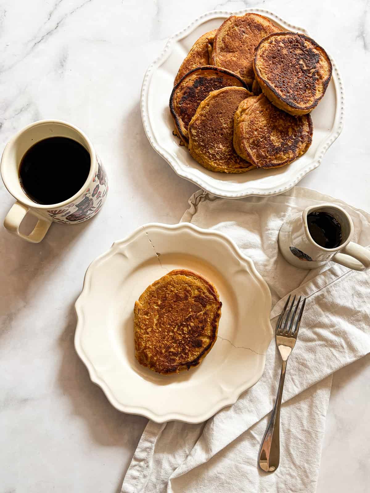 Soft and fluffy gluten free pumpkin pancakes are packed with plenty of fall flavor thanks to pumpkin puree, warming spices, and maple syrup! A pitcher of maple syrup and gluten free pumpkin pancakes and a cup of coffee.
