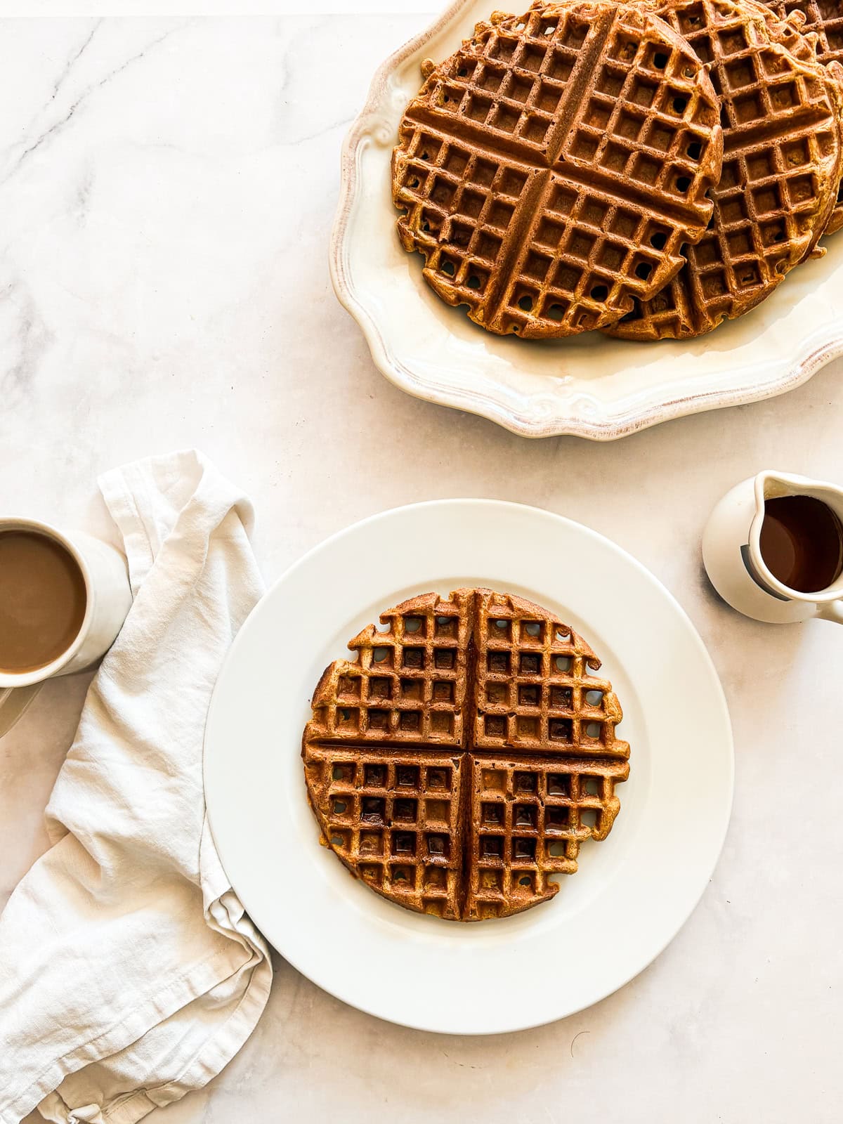 Warmly spiced and naturally gluten-free pumpkin waffles are what you want on a crisp fall morning. Made with whole grain oat flour! Pumpkin waffles on a plate and a serving platter of waffles.